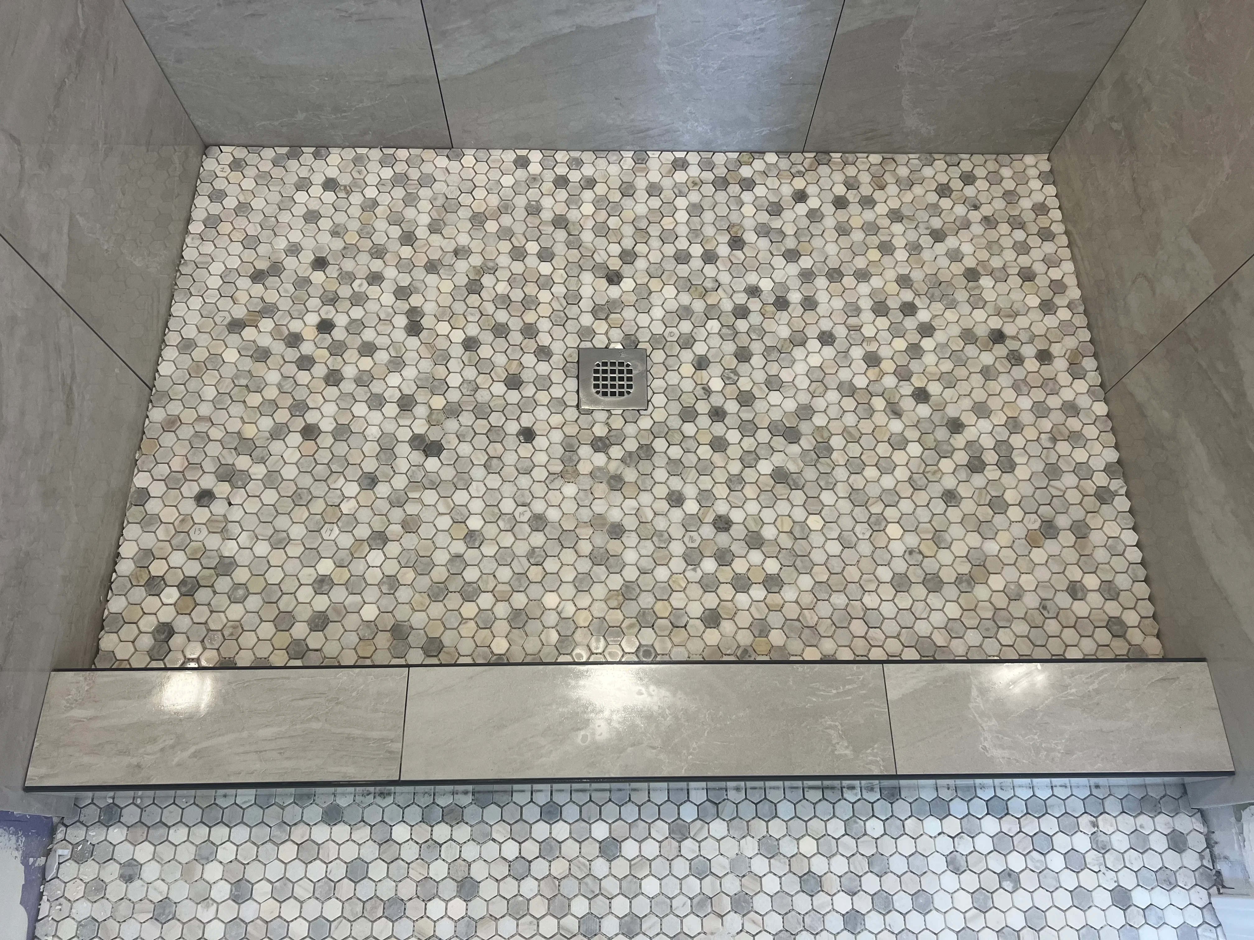 Hexagon mosaic tile shower floor with cream and gray tiles, centered drain, and concrete walls