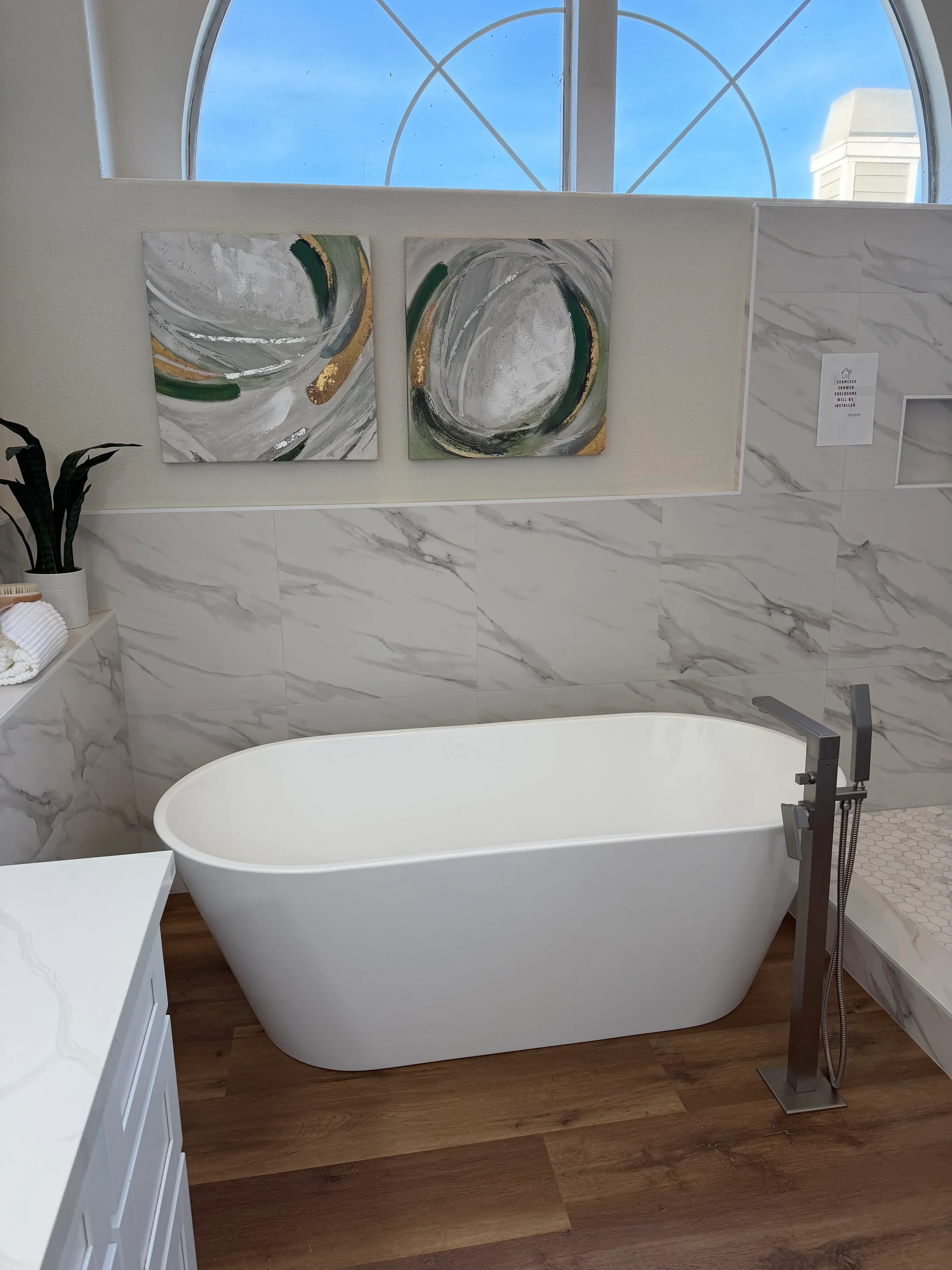 White oval sink with black faucet, gray marble tile walls, wood flooring, and decorative artwork